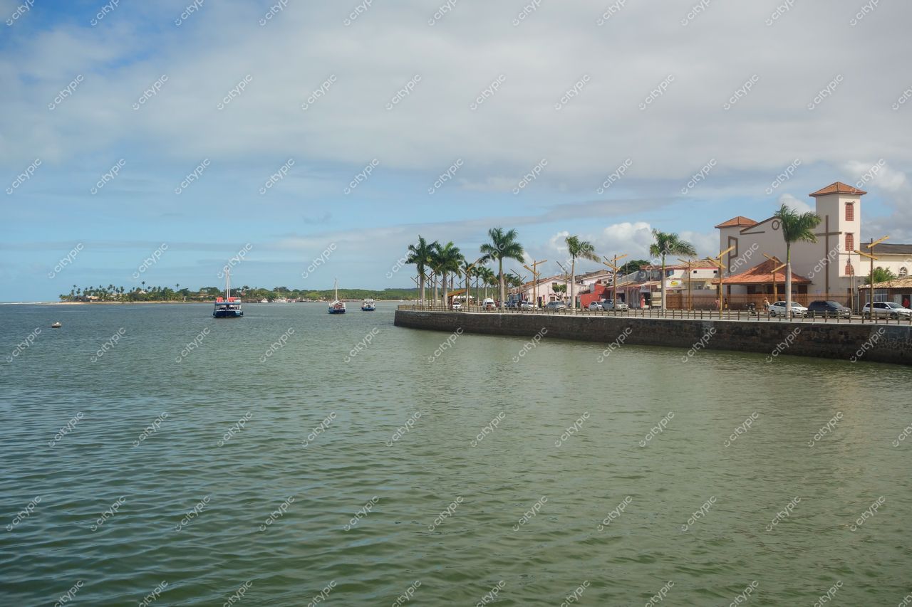 Porto Seguro, Bahia, orla com barcos ancorados e calçadão arborizado em dia ensolarado e céu parcialmente nublado