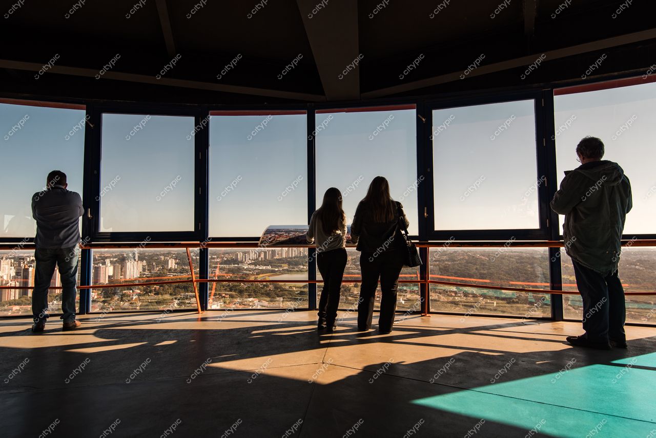 Curitiba, Paraná, pessoas observando a paisagem urbana através de janelas grandes com luz da tarde e sombras marcantes