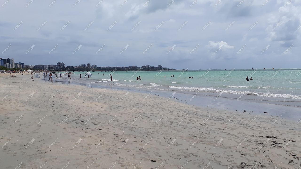 Maceió, Alagoas, praia com areia clara e úmida, céu nublado e edifícios à beira-mar ao fundo