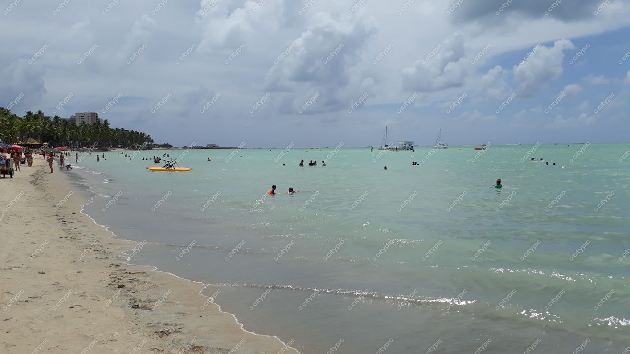 Maceió, Alagoas com águas calmas e esverdeadas, banhistas na praia e palmeiras ao fundo em dia nublado