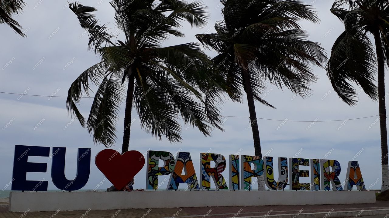 Paripueira, Alagoas com letreiro “EU ❤️ PARIPUEIRA” em calçada, palmeiras e céu nublado ao fundo