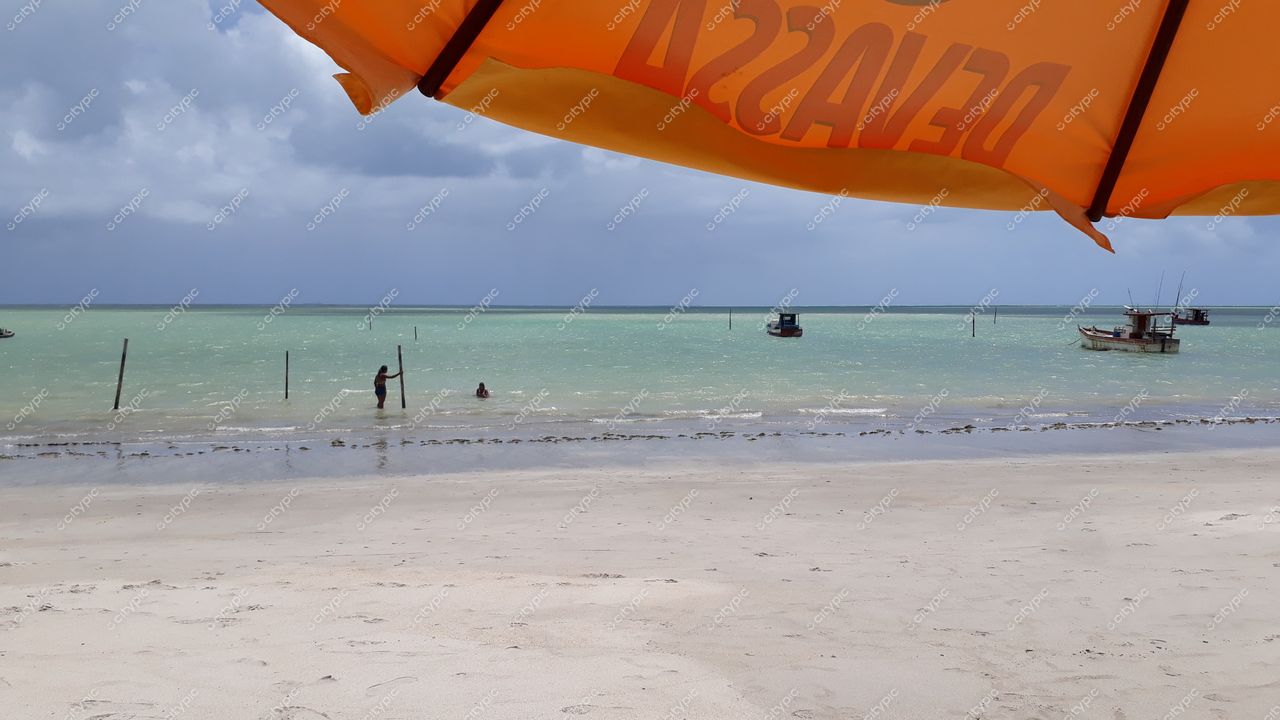 Japaratinga, Alagoas com guarda-sol laranja e pessoas na água clara em dia parcialmente nublado e embarcações ao fundo