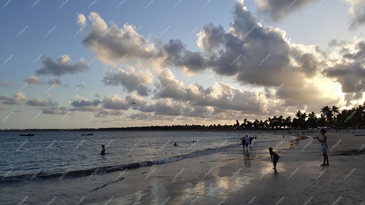 Porto de Galinhas, Pernambuco, entardecer com céu nublado e pessoas na areia entre coqueiros e embarcações