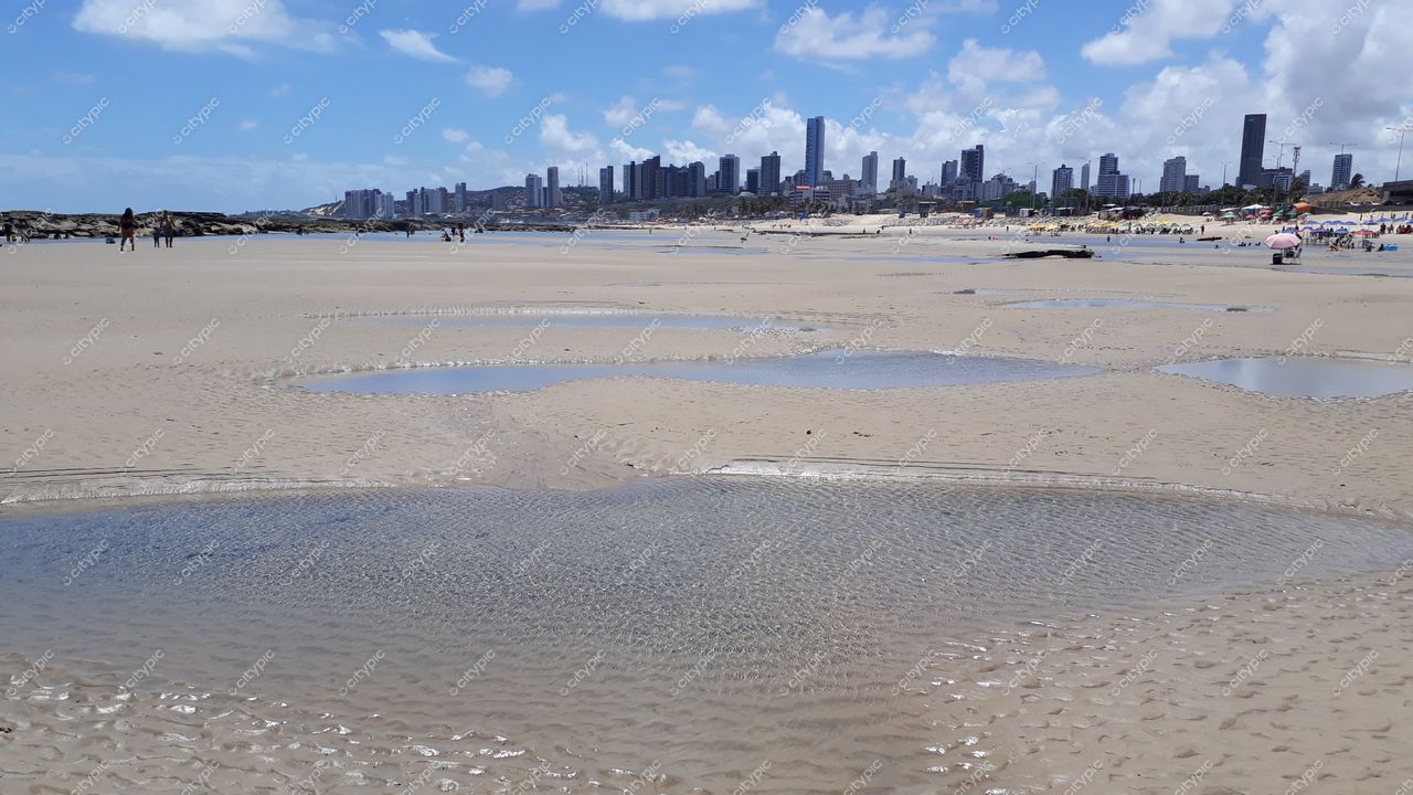 Natal, Rio Grande do Norte com praia ensolarada, céu parcialmente nublado e orla urbana ao fundo com pessoas caminhando