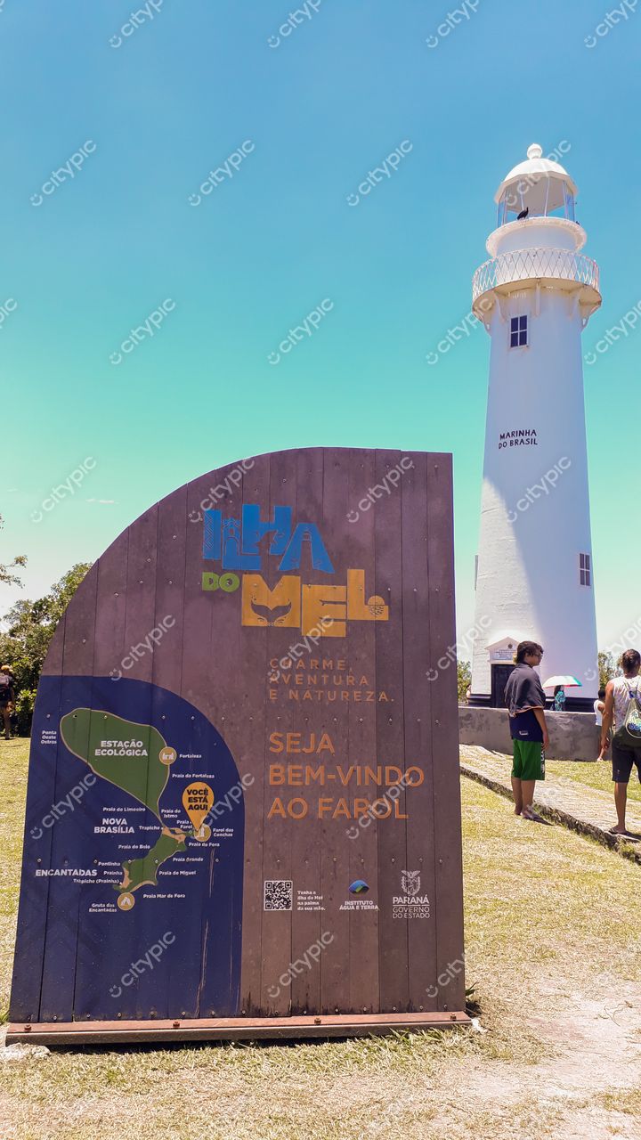 Ilha do Mel, Paraná, painel de madeira curvo com informações turísticas e farol branco sob céu ensolarado