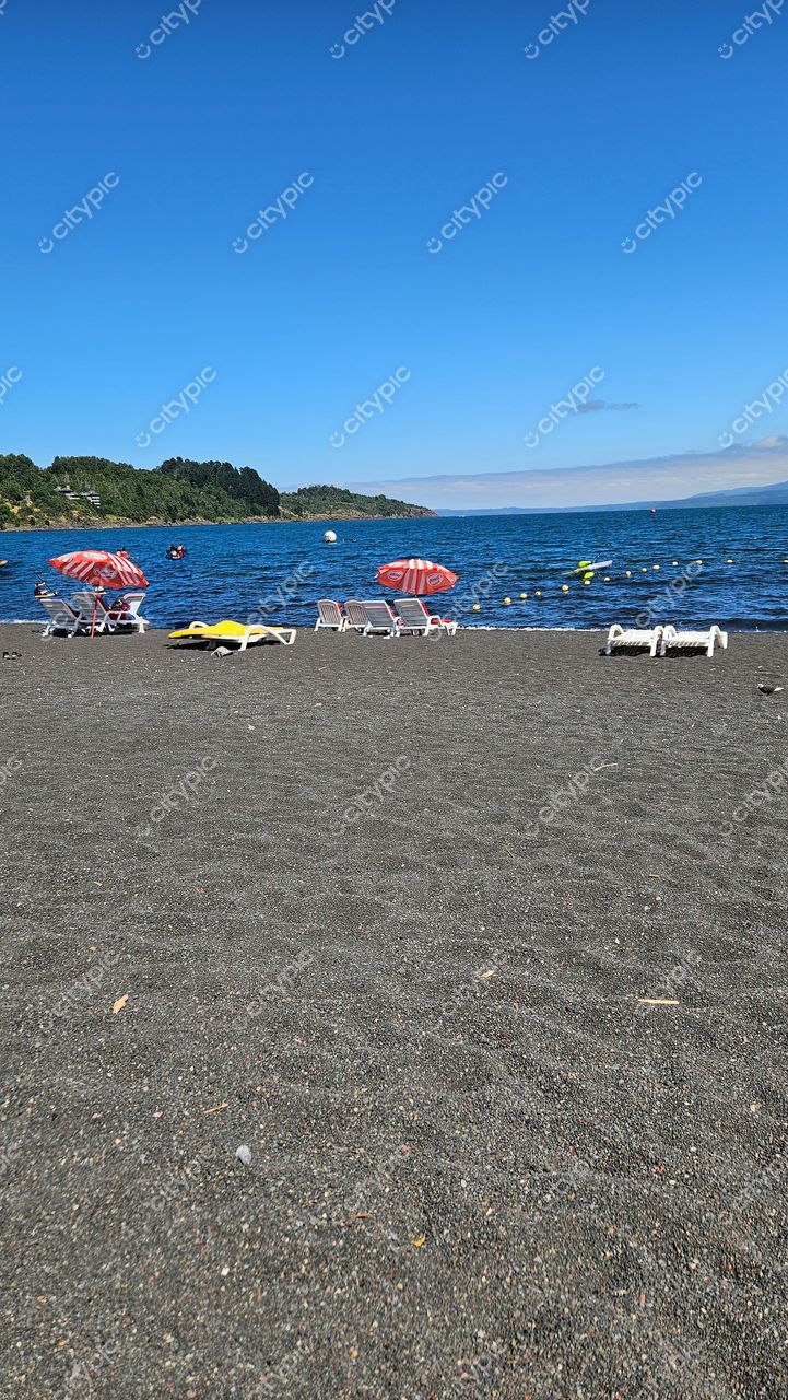 Praia de areia escura com cadeiras brancas e guarda-sóis listrados, lago calmo e vegetação verde ao fundo