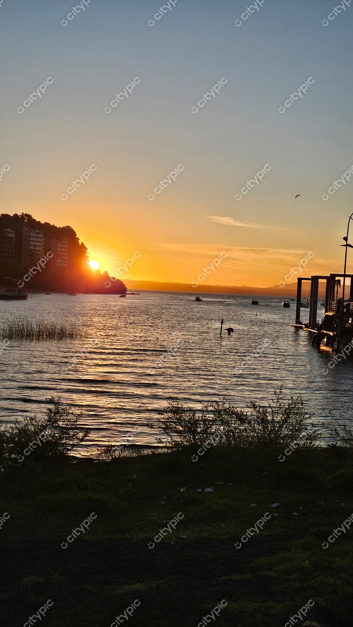 Pôr do sol à beira de lago com reflexos dourados, silhuetas de prédios e vegetação densa ao fundo