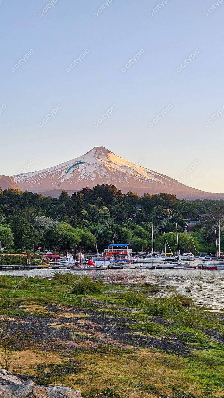 Cenário natural ao entardecer com montanha nevada, corpo d’água e barcos ancorados em pequeno porto