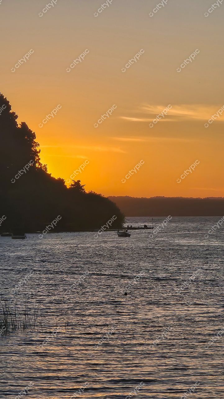 Pôr do sol sereno sobre lago calmo com reflexos quentes e silhueta de vegetação densa à esquerda