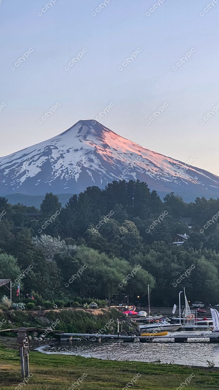 Vulcão coberto de neve ao pôr do sol com marina e vegetação densa em cenário montanhoso tranquilo