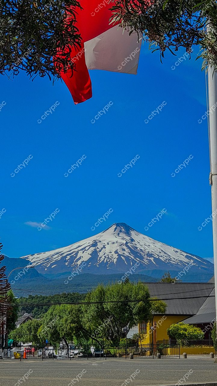 Cenário montanhoso com vulcão nevado ao fundo e bandeira vermelha em meio à vegetação rural tranquila
