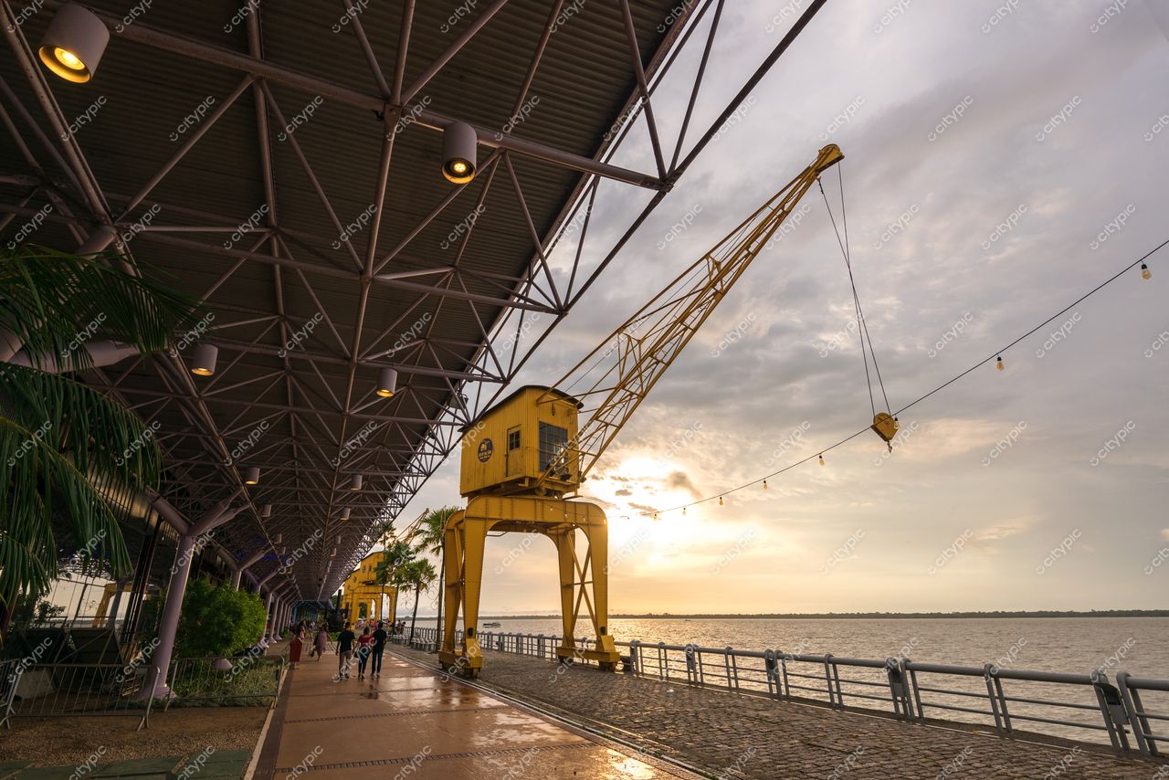 Belém, Pará, guindaste amarelo sob teto metálico ao entardecer com pessoas no calçadão e vegetação tropical