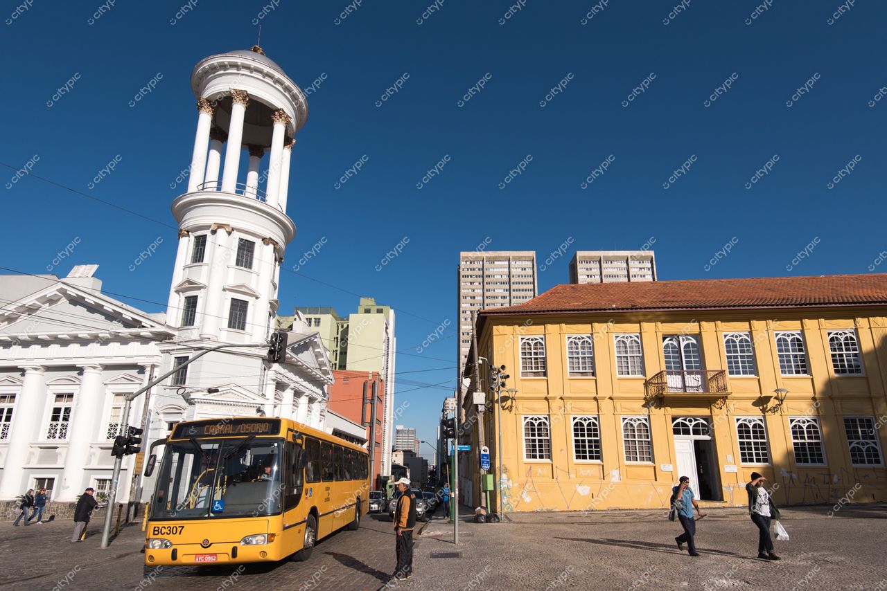 Curitiba, Paraná, ônibus amarelo em destaque com edifícios coloniais brancos e amarelos sob céu azul ensolarado