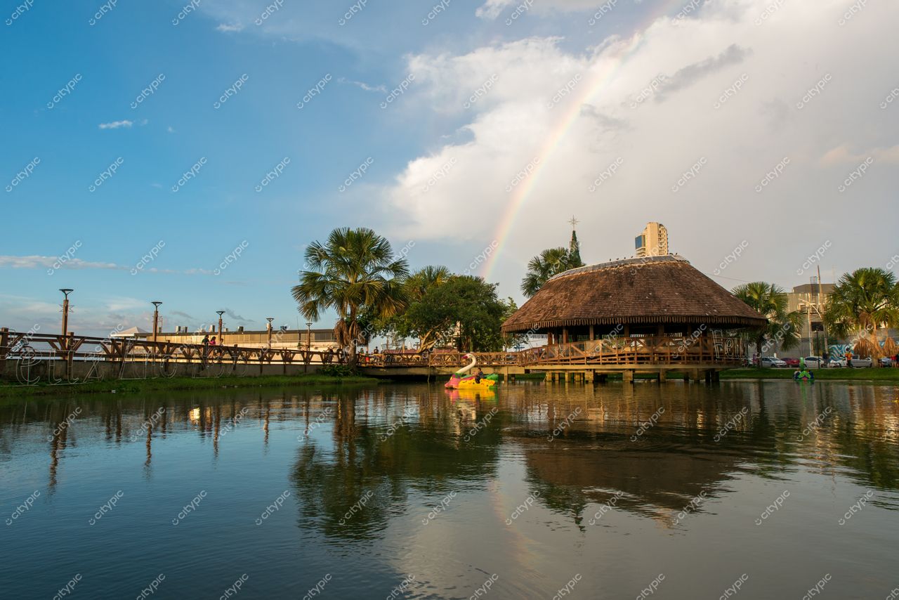 Belém, Pará, lago tranquilo ao entardecer com reflexos e construção de palha sob arco-íris e vegetação típica