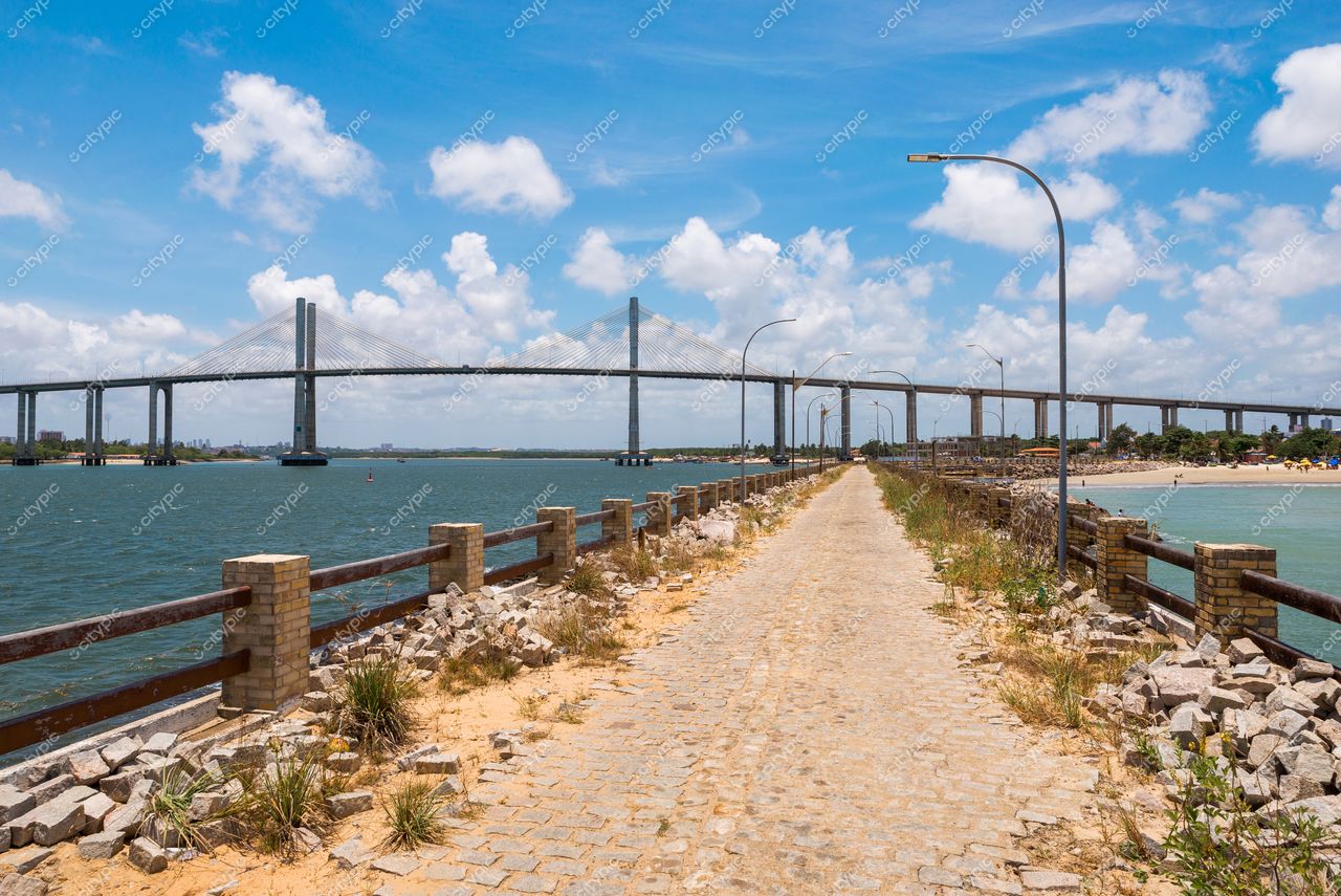 Natal, Rio Grande do Norte, vista do litoral com calçamento, vegetação lateral e ponte ao fundo sob céu ensolarado