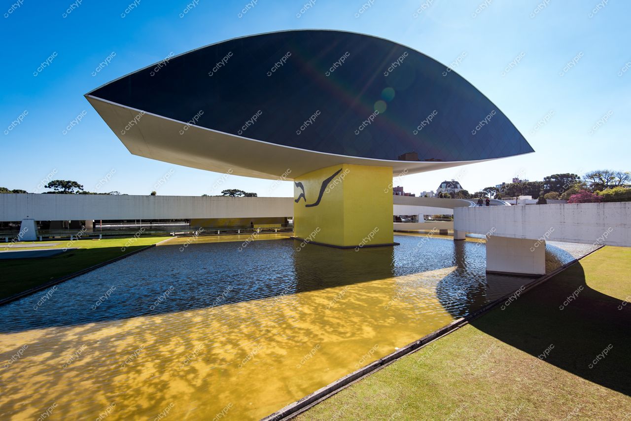 Curitiba, Paraná Museu Oscar Niemeyer refletido em água tranquila com vegetação ao fundo e céu limpo durante o dia