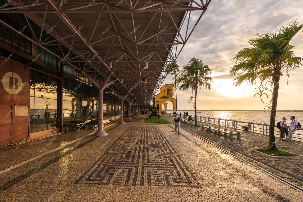 Belém, Pará, calçada de pedras portuguesas sob teto metálico com palmeiras e rio ao fundo no pôr do sol