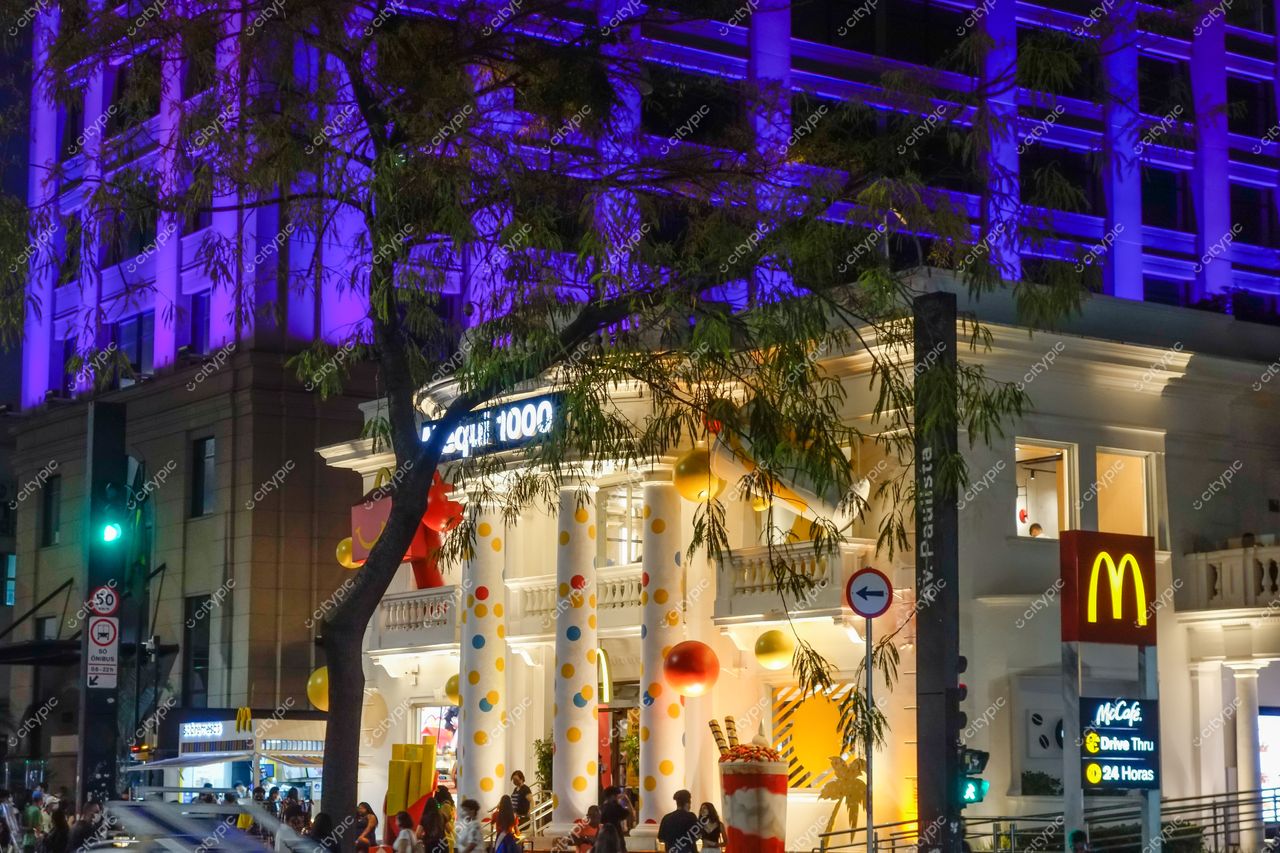 São Paulo, São Paulo, esquina movimentada com McDonald’s decorado, prédio roxo ao fundo e iluminação noturna vibrante