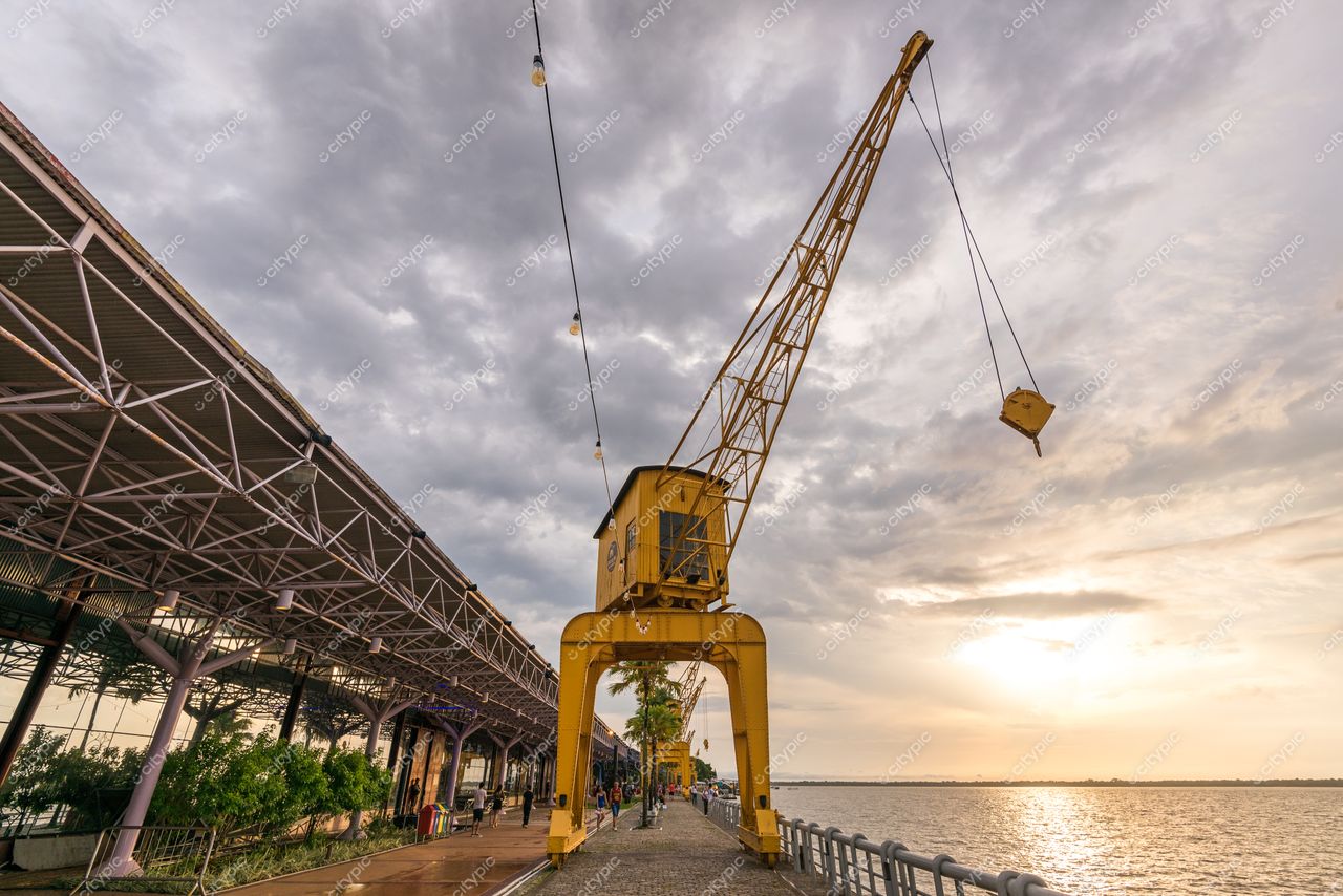 Belém, Pará com guindaste amarelo à beira do rio em entardecer nublado e vegetação urbana ao fundo