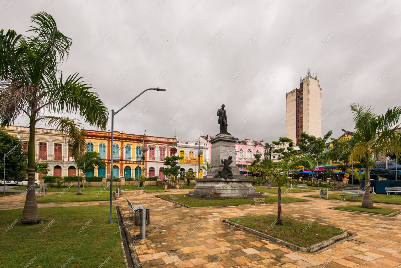 Belém, Pará, praça com monumento central rodeado por gramado e palmeiras em dia nublado com edifícios coloridos ao fundo