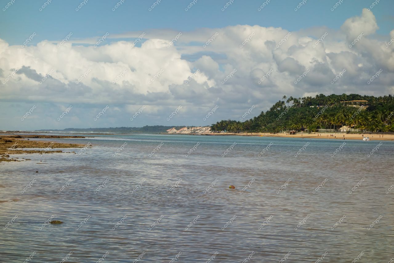 Arraial D’ajuda, Bahia com água rasa refletindo céu azul e nuvens, vegetação tropical e formações rochosas à vista