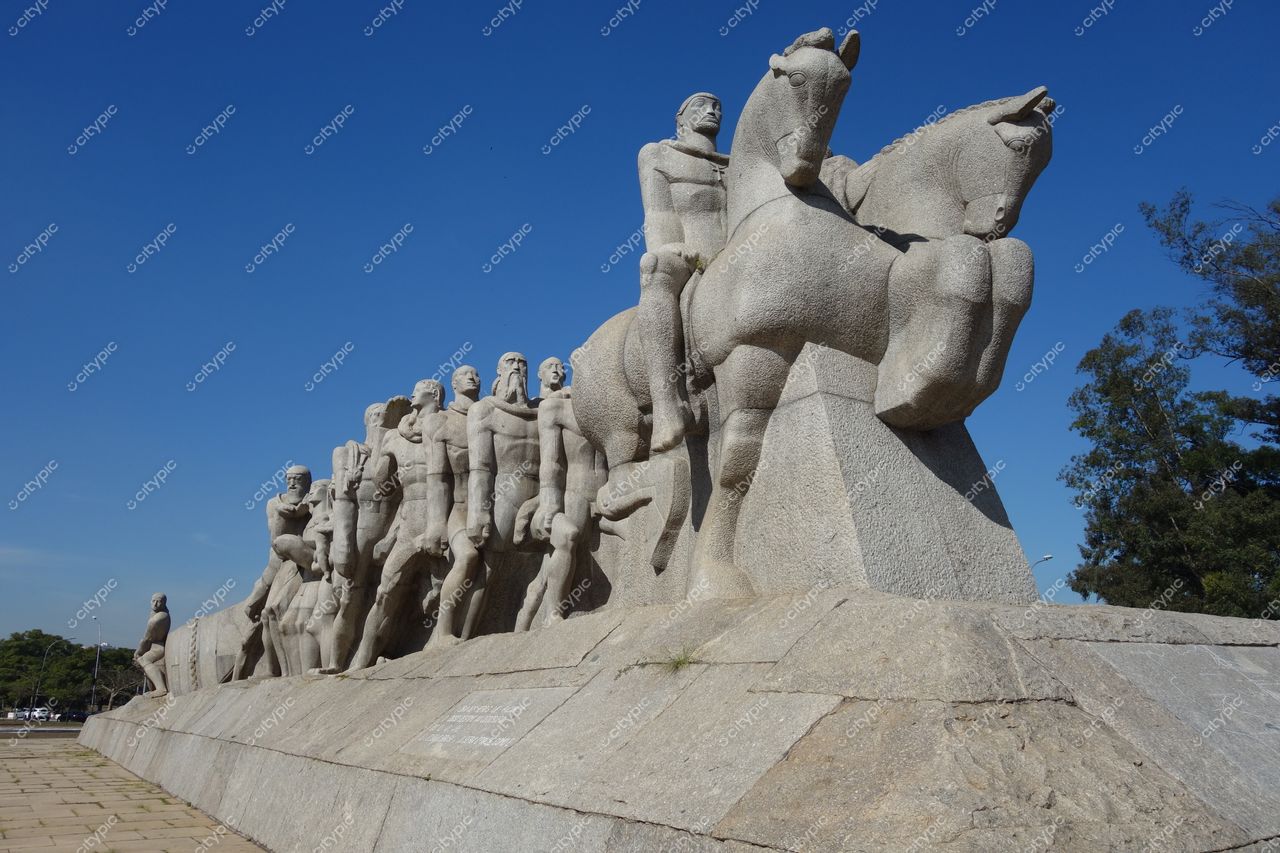 Monumento às Bandeiras em São Paulo com céu azul e vegetação ao redor, destacando a escultura de granito e a paisagem urbana