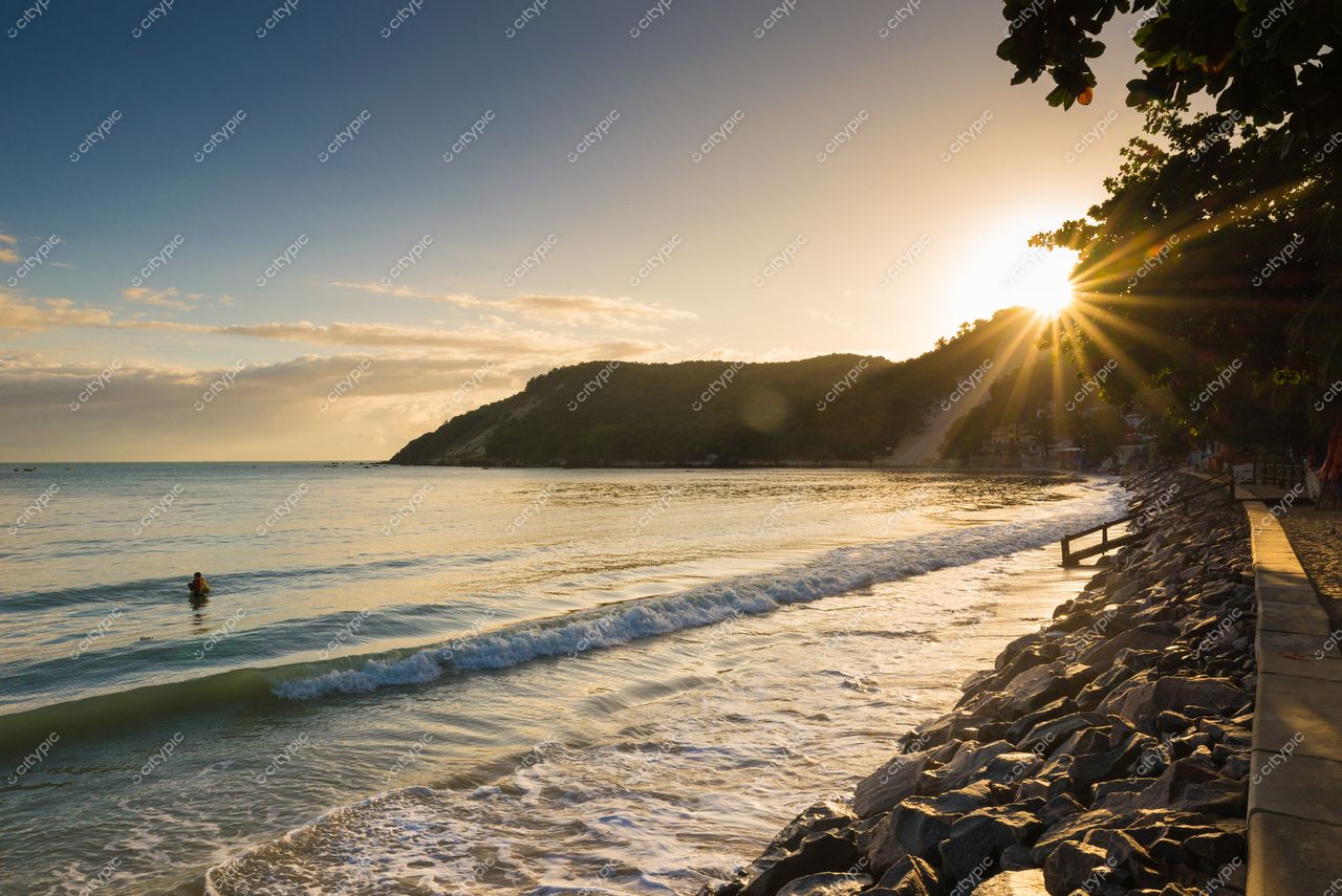 Natal, Rio Grande do Norte, pôr do sol na praia com ondas suaves e morro coberto de vegetação ao fundo
