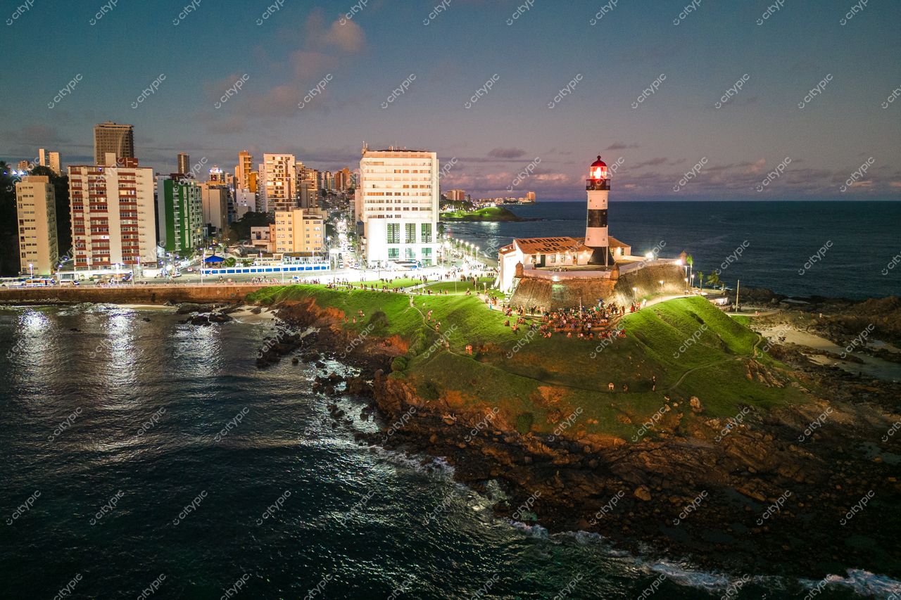Salvador, Bahia, cenário noturno com o Farol da Barra iluminado e reflexos na baía tranquila e urbana