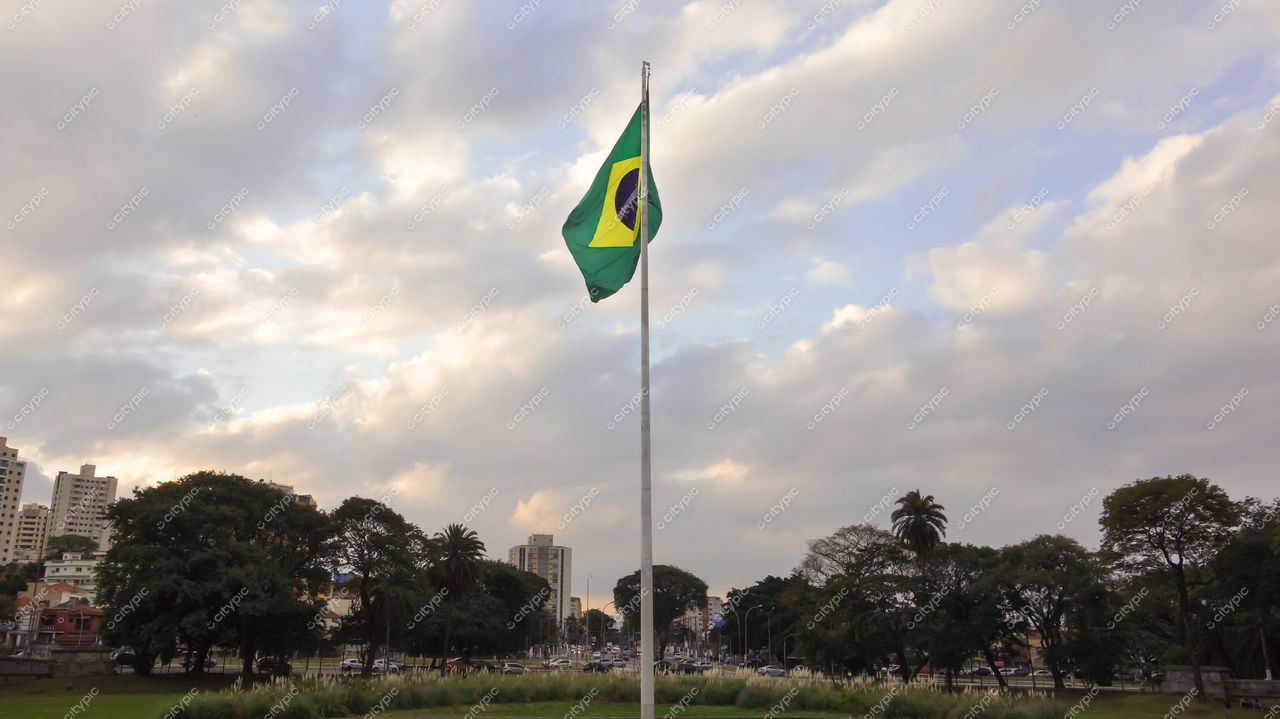 São Paulo, São Paulo, bandeira do Brasil hasteada em área verde com edifícios ao fundo e céu de fim de tarde