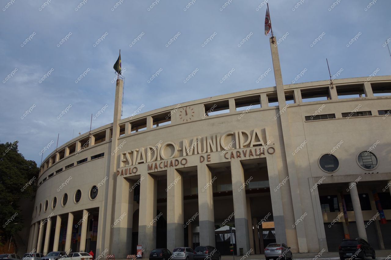 Estádio do Pacaembu, em São Paulo, com fachada curvada e bandeiras ao entardecer entre vegetação e veículos