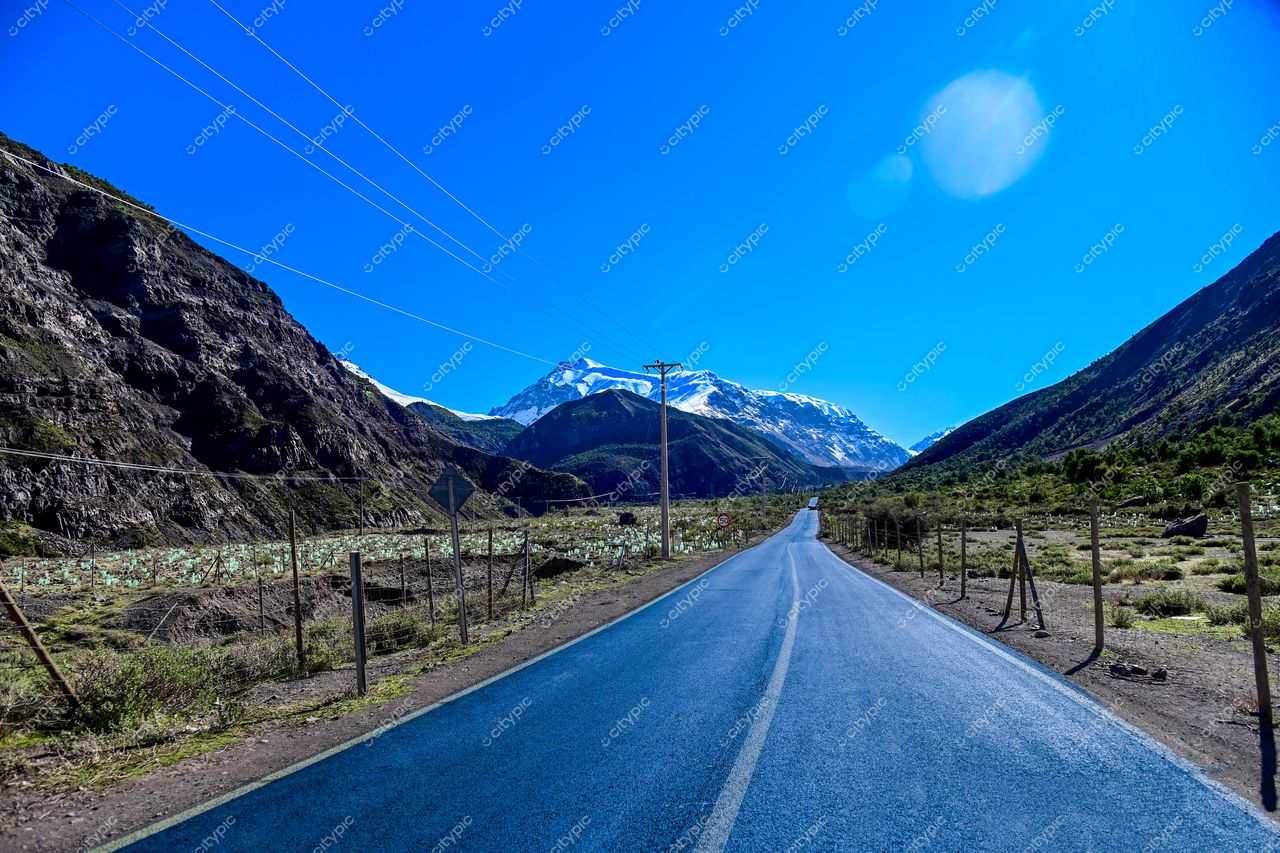 Estrada asfaltada em direção a montanhas nevadas sob céu azul, cercada por formações rochosas e campos cultivados