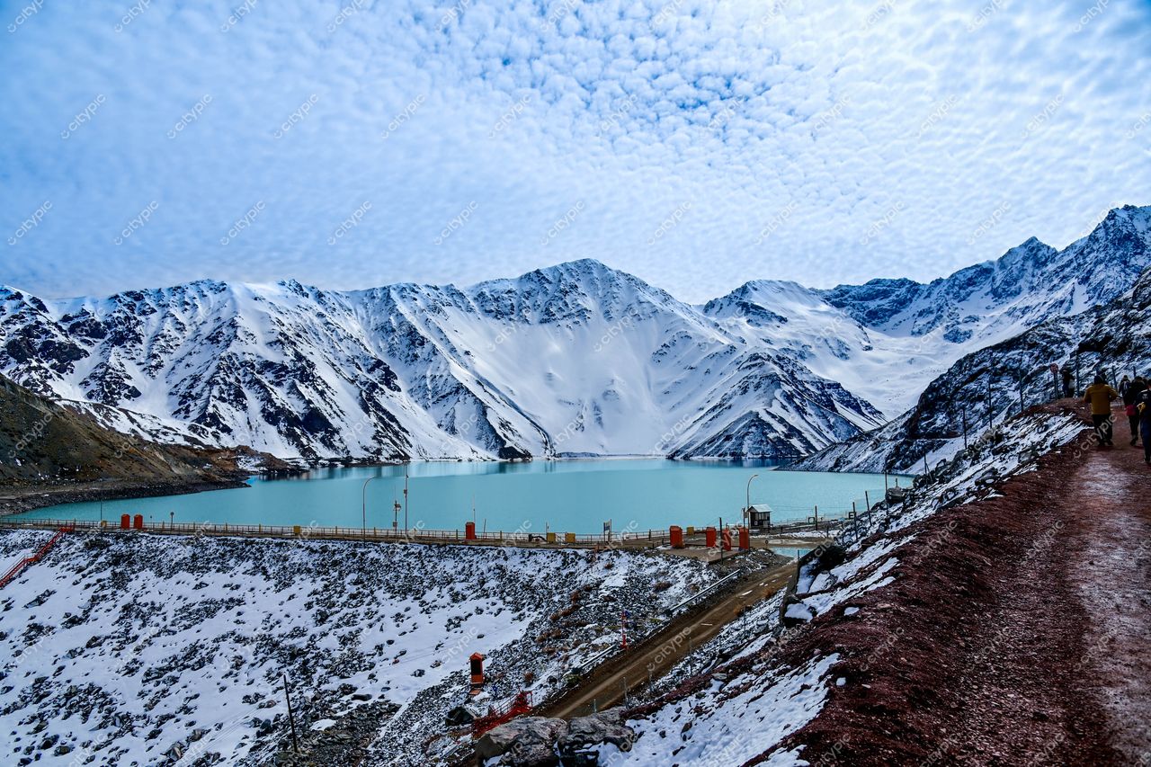 Paisagem montanhosa com picos nevados e lago azul-turquesa, estrada ao longo da margem e nuvens no céu