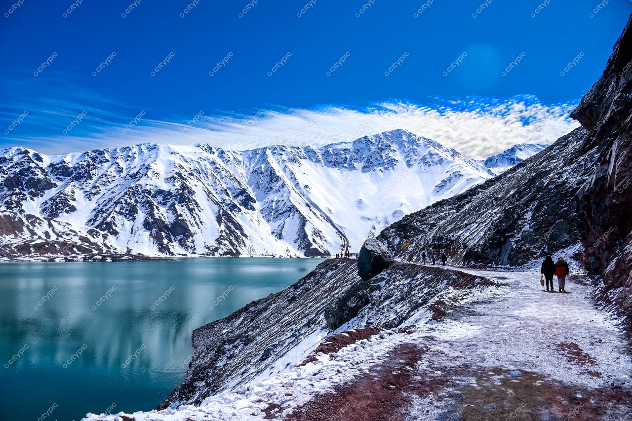 Caminho estreito ao longo de lago azul em paisagem montanhosa com picos nevados e céu ensolarado