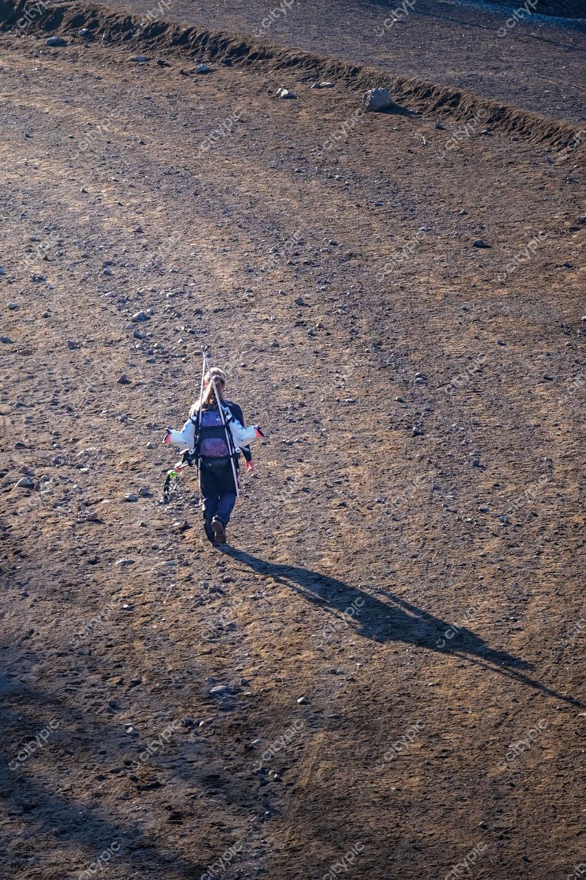 Figura humana caminhando em terreno árido sob luz suave do sol baixo, carregando equipamento colorido nas costas
