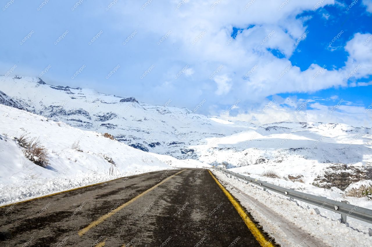 Estrada sinuosa coberta de neve em região montanhosa com picos brancos e céu parcialmente nublado iluminando a cena