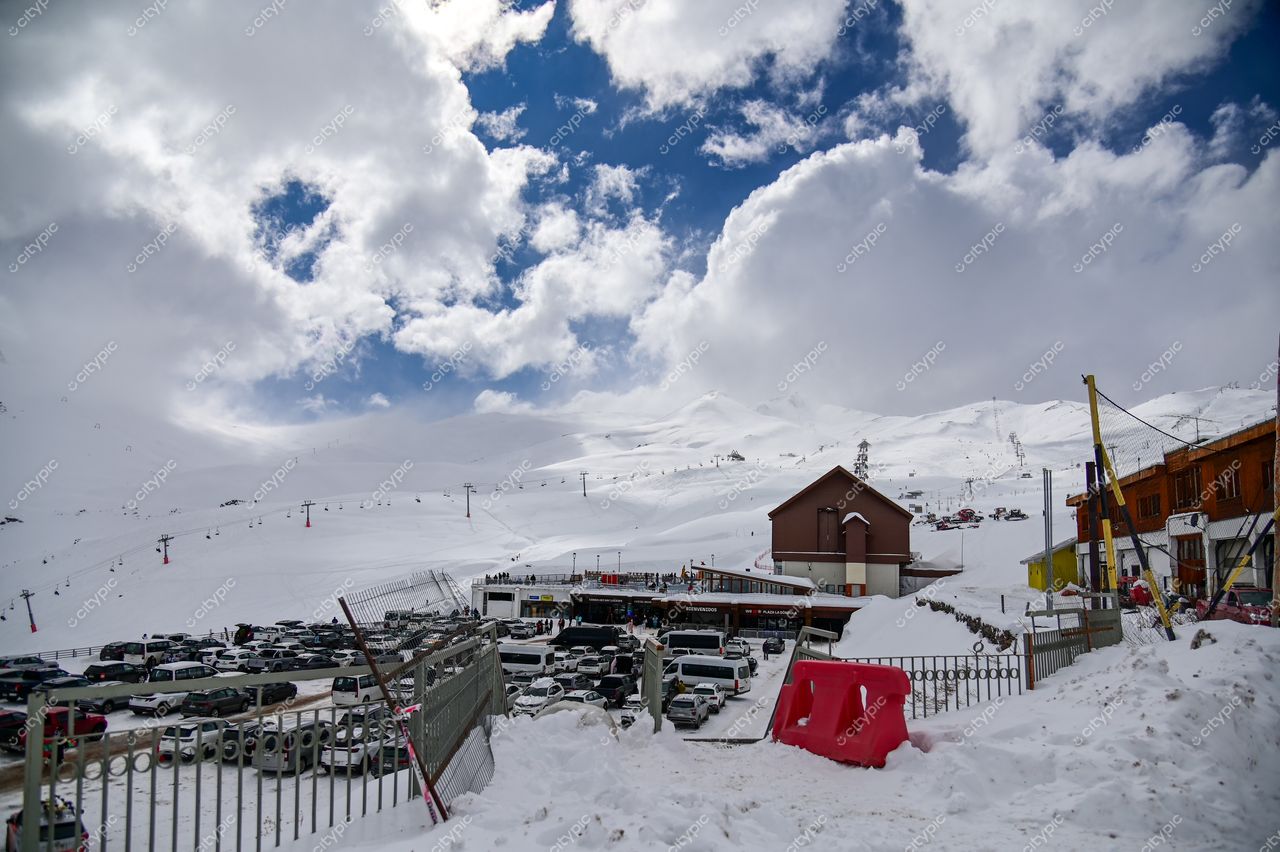 Estacionamento cheio em área montanhosa nevada com teleféricos e edifícios de madeira sob céu parcialmente nublado