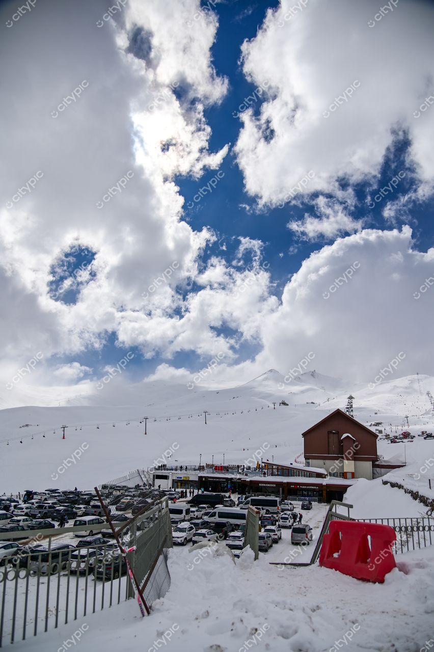 Estação de esqui com estacionamento cheio e visitantes em dia nublado, montanha ao fundo e teleféricos visíveis