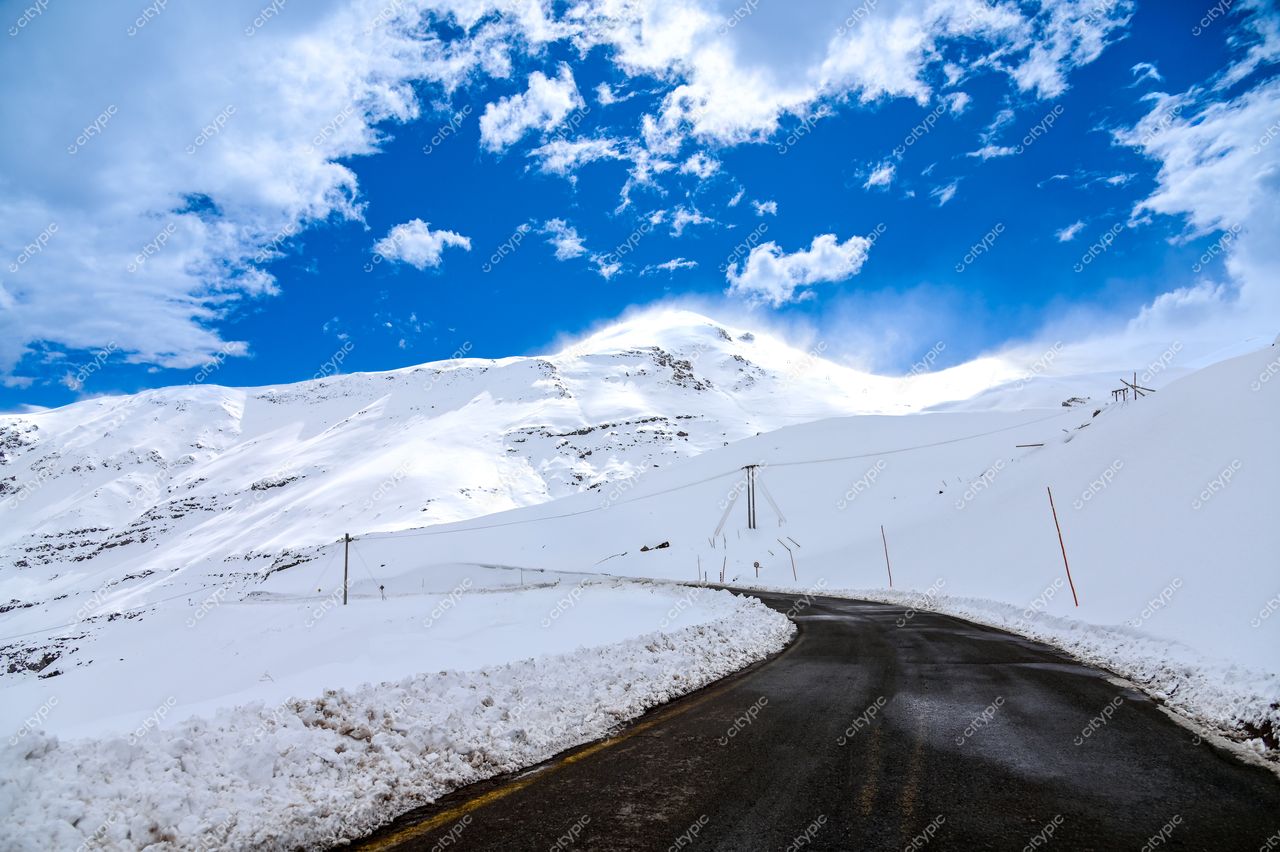 Estrada sinuosa coberta de neve em paisagem montanhosa sob céu azul claro com nuvens brancas esparsas