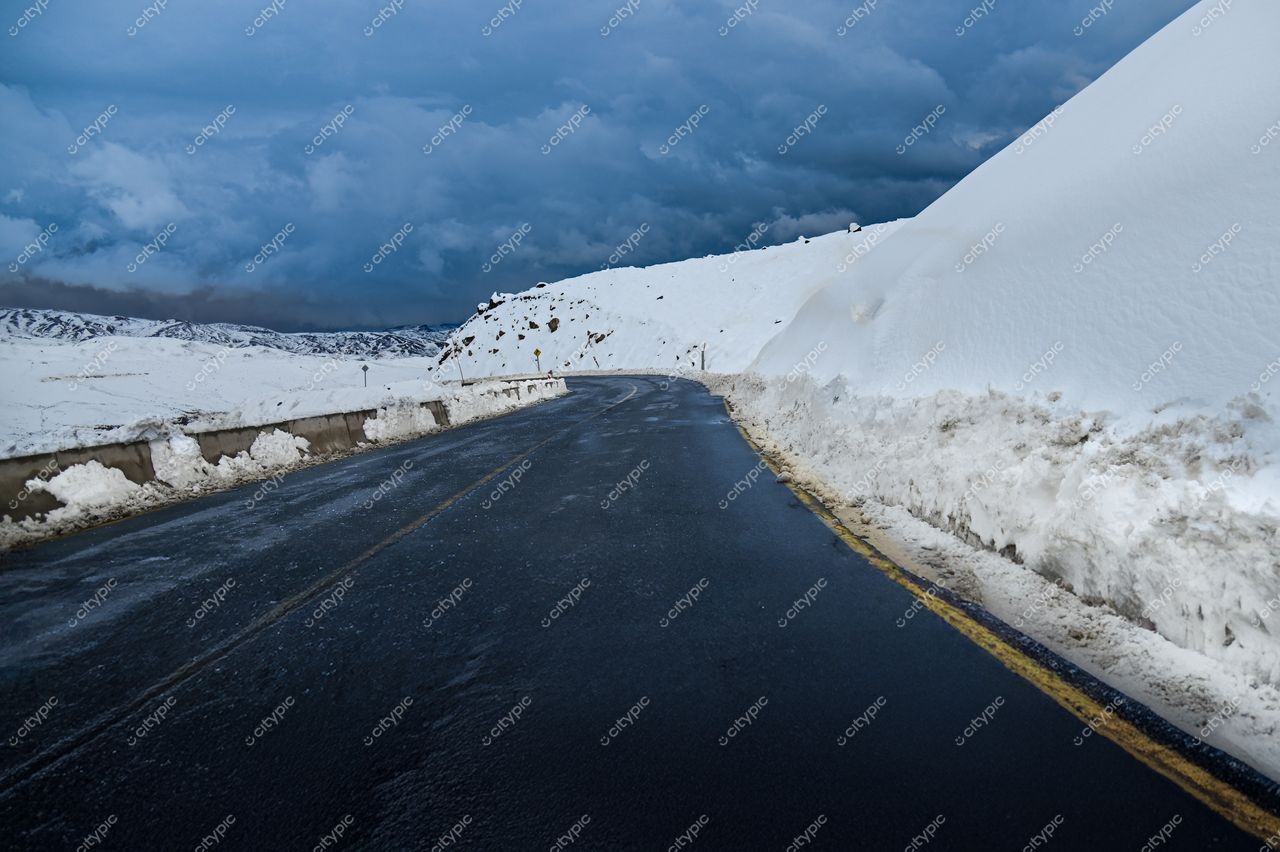 Estrada asfaltada em ambiente invernal com montanhas nevadas e céu nublado, sem tráfego ou vegetação visível