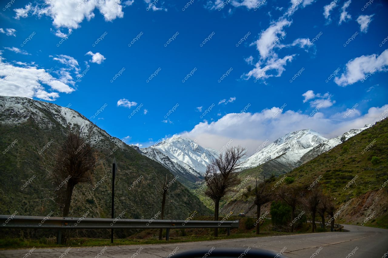 Paisagem montanhosa com picos nevados e céu azul, árvores desfolhadas ao longo de estrada sinuosa em dia ensolarado