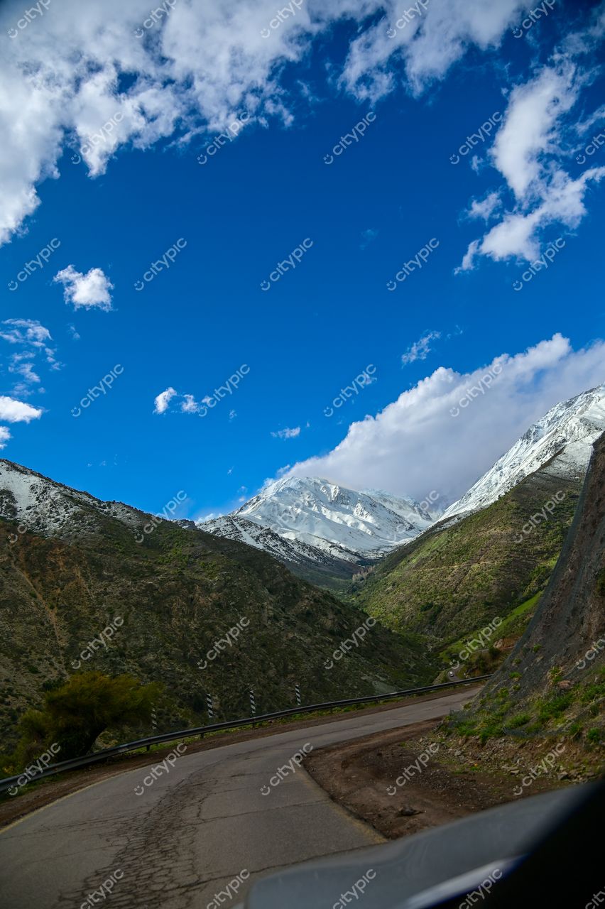 Estrada sinuosa em paisagem montanhosa com pico nevado ao fundo sob céu azul e nuvens esparsas