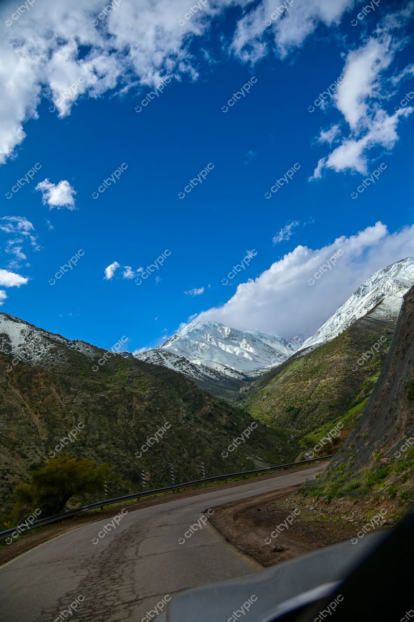 Estrada sinuosa em montanha verdejante com picos nevados ao fundo sob céu azul e nuvens brancas