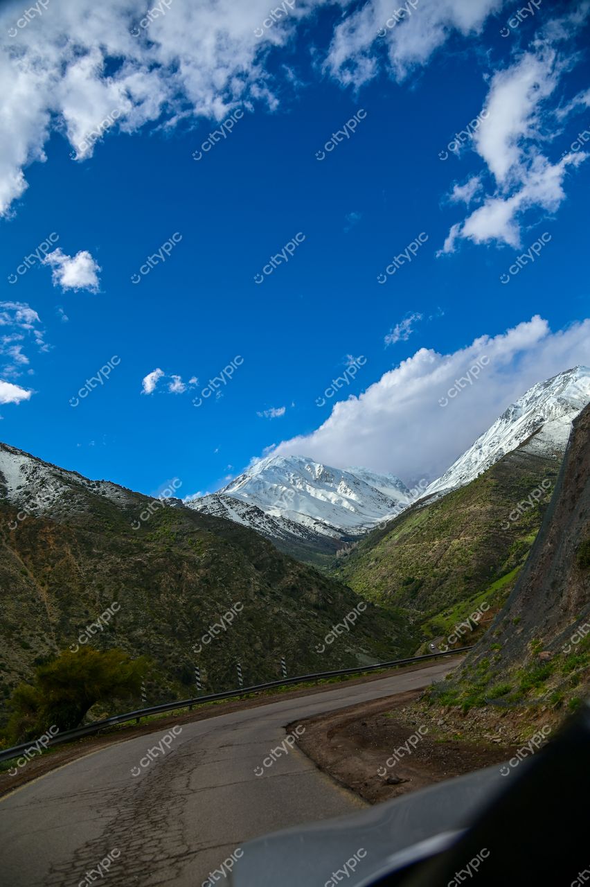 Estrada sinuosa em vale montanhoso com vegetação verde e montanhas nevadas sob céu azul durante o dia