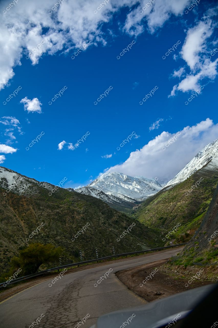 Estrada sinuosa em montanha com vegetação à esquerda e pico nevado à direita sob céu parcialmente nublado