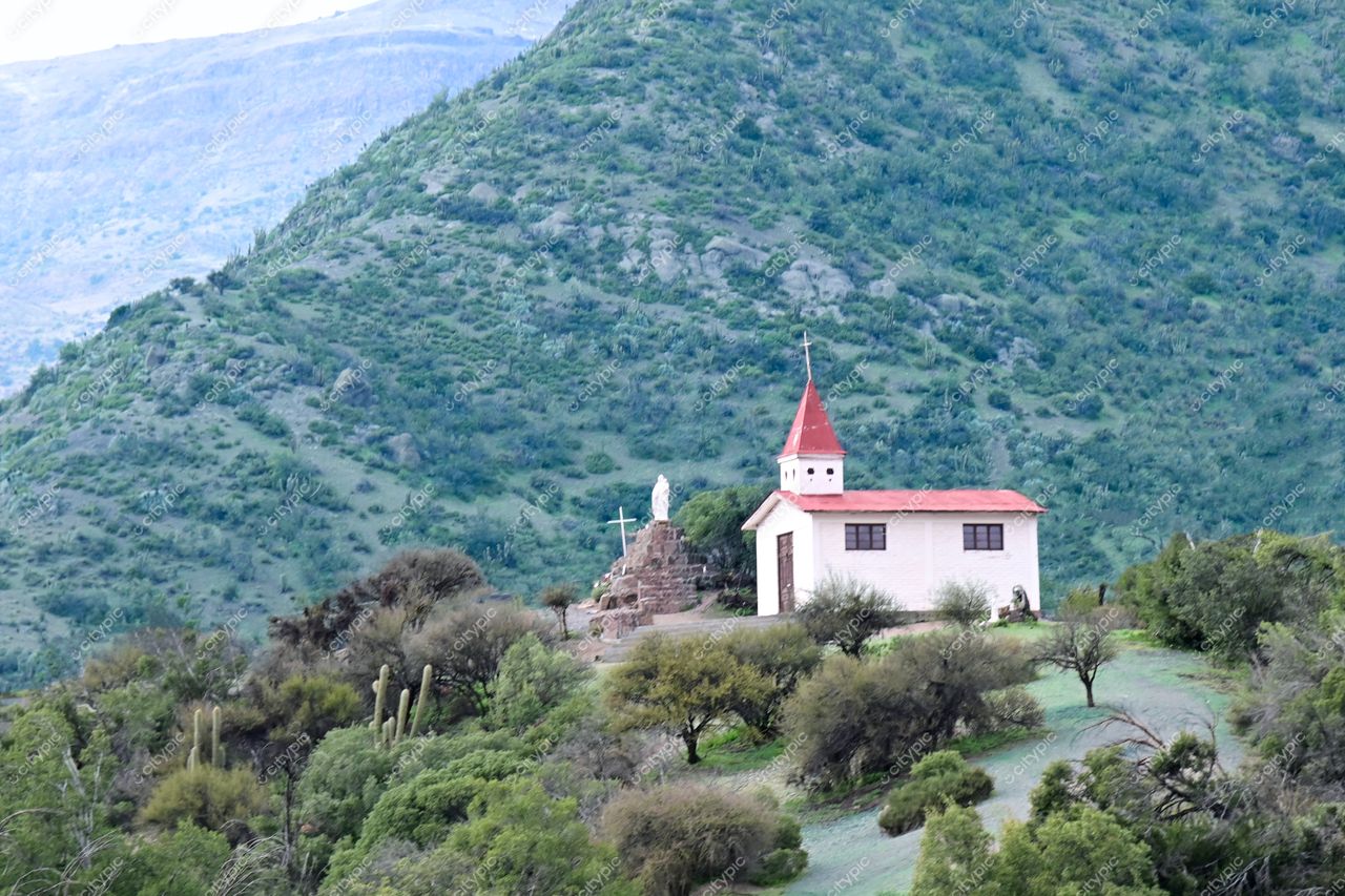 Igreja branca com telhado vermelho em encosta montanhosa sob céu nublado, cercada por vegetação nativa e estrutura de pedra