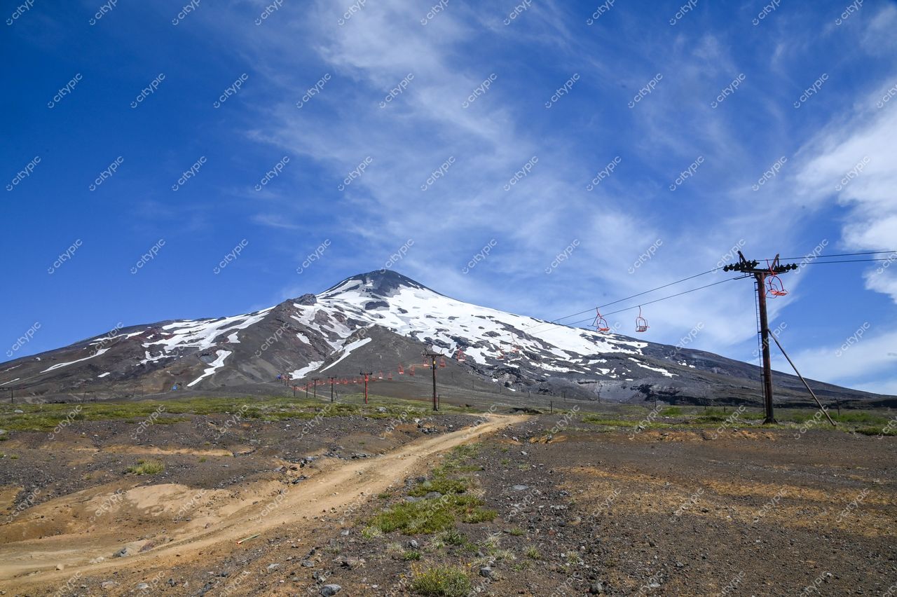 Vulcão coberto de neve com relevo acidentado e estrada de terra sob céu azul claro e vegetação esparsa