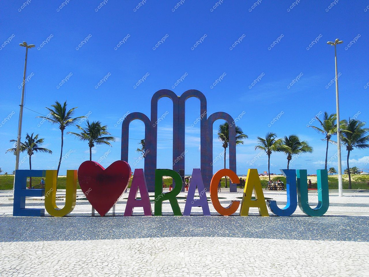 Letreiro Eu Amo Aracaju em Aracaju com céu azul, palmeiras ao fundo e calçamento em pedras urbanas