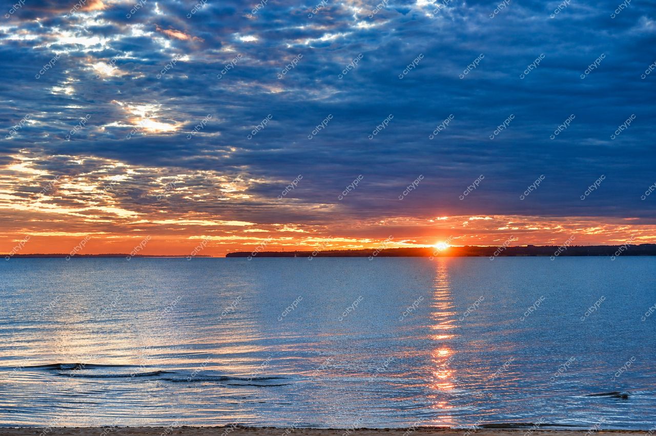 Pôr do sol sereno sobre corpo d’água calmo com reflexos quentes e praia de areia ao primeiro plano