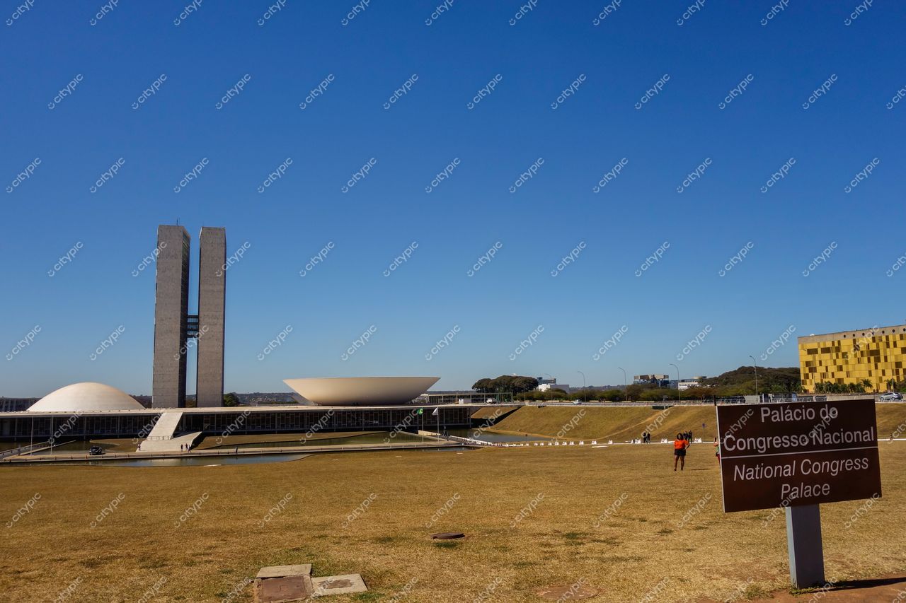 Congresso Nacional em Brasília com torres verticais e cúpulas sob céu azul e gramado, sinal indicativo em primeiro plano