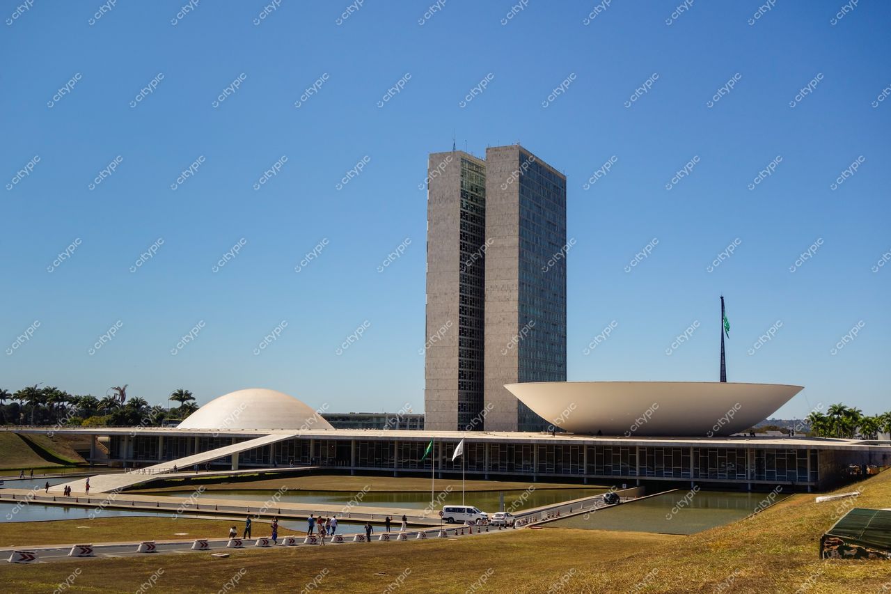 Congresso Nacional em Brasília com luz do dia, céu claro, vegetação ao fundo e pessoas no espaço aberto